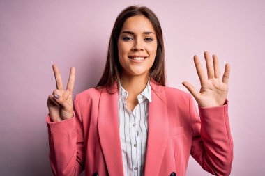 Güzel, esmer, ceketli bir kadın pembe arka planda duruyor. Yedi numaralı parmaklarıyla işaret ediyor ve gülümsüyor. Kendinden emin ve mutlu..