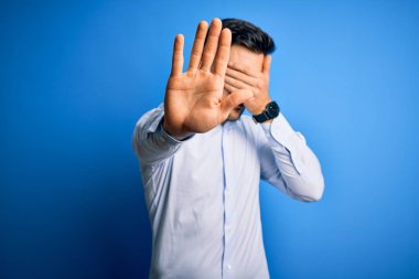 Young handsome man wearing elegant shirt standing over isolated blue background covering eyes with hands and doing stop gesture with sad and fear expression. Embarrassed and negative concept.