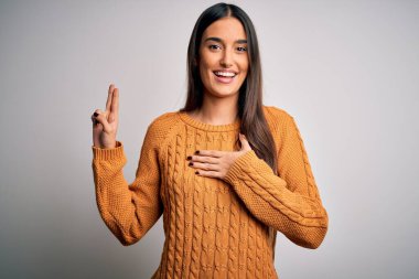 Genç, güzel esmer kadın, izole edilmiş beyaz arka plan üzerine gündelik süveter giyip gülümsüyor, elleri göğsünde ve parmakları havada küfür ediyor, sadakat yemini ediyor.