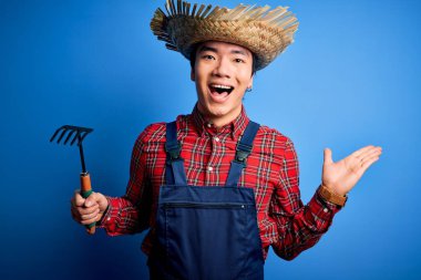Young handsome chinese farmer man wearing apron and straw hat holding rake tool very happy and excited, winner expression celebrating victory screaming with big smile and raised hands