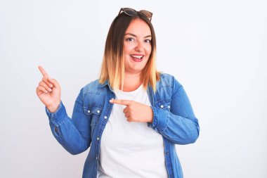 Güzel bir kadın kot gömlek giyiyor, beyaz arka planda duruyor gülümsüyor ve kameraya bakıyor, iki eliyle ve parmaklarıyla kenarı işaret ediyor..