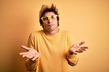 Sıradan tişört ve gözlük takan genç yakışıklı bir adam izole edilmiş sarı arka plan bilgisiz ve elleri ve kolları havada kafası karışmış bir ifade takınıyor. Şüphe kavramı.