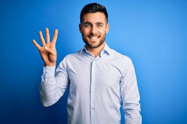 Zarif gömlekli genç adam mavi arka plan üzerinde duruyor ve dört numaralı parmaklarıyla işaret ederken kendinden emin ve mutlu bir şekilde gülümsüyor..