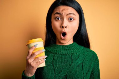 Genç, güzel Çinli bir kadın izole edilmiş sarı arka planda kahve içiyor. Şaşırmış bir yüzle korkmuş ve korku ifadesinden heyecanlanmış.