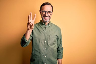 Middle age hoary man wearing casual green shirt and glasses over isolated yellow background showing and pointing up with fingers number three while smiling confident and happy.