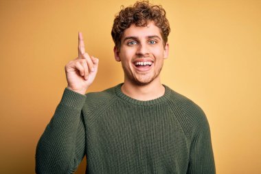Young blond handsome man with curly hair wearing green sweater over yellow background pointing finger up with successful idea. Exited and happy. Number one.
