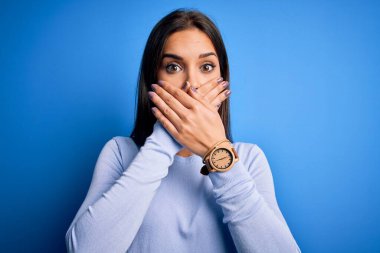 Young beautiful brunette woman wearing casual sweater standing over blue background shocked covering mouth with hands for mistake. Secret concept.