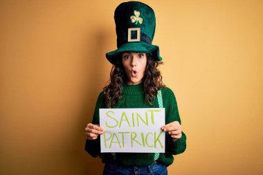 Beautiful curly hair woman wearing green hat holding banner with saint patricks message scared in shock with a surprise face, afraid and excited with fear expression