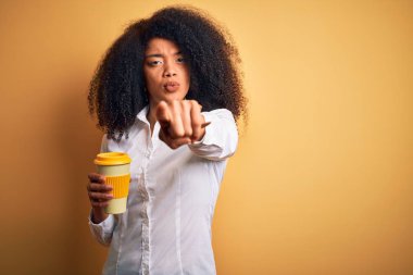 Afro saçlı, kahve fincanından kahve içen, kamerayı işaret eden Afro-Amerikalı genç bir iş kadını ve sen, el işareti, ön taraftan pozitif ve kendinden emin bir jest.