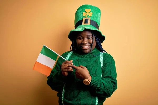 非洲裔美国妇女戴着绿色的帽子 在圣帕特里克日手持爱尔兰国旗 她非常高兴地用手和手指指指点点 — 图库照片