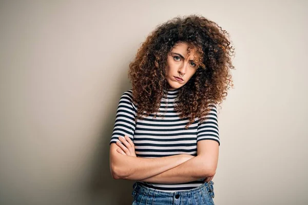 Young beautiful woman with curly hair and piercing wearing casual striped t-shirt skeptic and nervous, disapproving expression on face with crossed arms. Negative person.