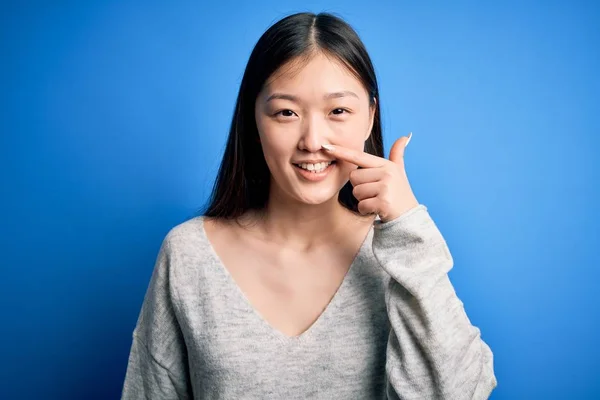 青い隔離された背景の上に立ってカジュアルなセーターを着て若い美しいアジアの女性顔と鼻に手の指で指して 陽気に笑顔 美の概念 — ストック写真