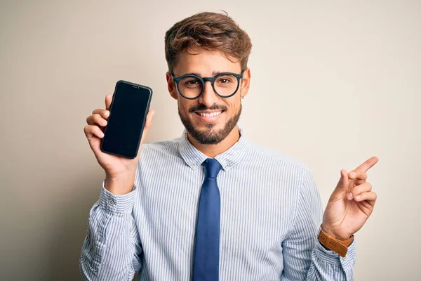 Akıllı telefonu elinde tutan genç bir işadamı izole edilmiş beyaz arka planda çok mutlu bir şekilde el ve parmak kenarı işaret ediyor.