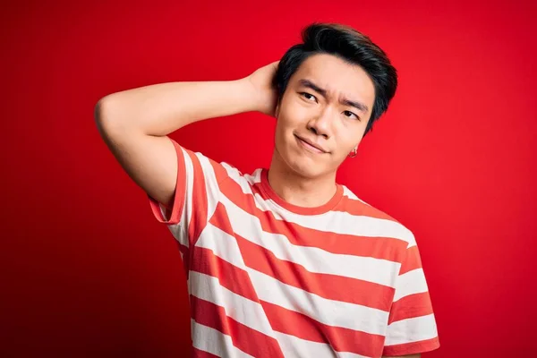 Young handsome chinese man wearing casual striped t-shirt standing over red background confuse and wondering about question. Uncertain with doubt, thinking with hand on head. Pensive concept.