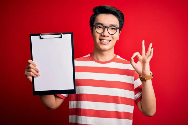 Young handsome chinese inspector man wearing glasses holding clipboard checklist doing ok sign with fingers, excellent symbol