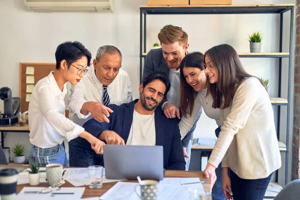 Group of business workers smiling happy and confident. One of them sitting and partners standing around. Working together with smile on face looking at the laptop at the office