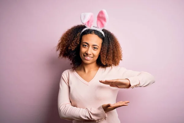 Young african american woman with afro hair wearing bunny ears over pink background gesturing with hands showing big and large size sign, measure symbol. Smiling looking at the camera. Measuring concept.