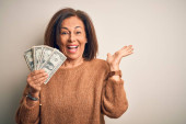 Středního věku brunetka žena drží jeden dolar bankovky na izolovaném pozadí velmi šťastný a vzrušený, vítěz výraz slaví vítězství křičí s velkým úsměvem a zvedl ruce