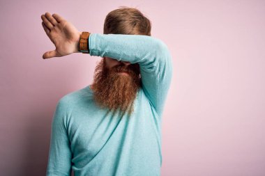 Sakallı, pembe izole edilmiş arka planda gözlüklü, yakışıklı kızıl saçlı, gözleri kollarıyla kaplayan, ciddi ve üzgün görünen bir adam. Görme, saklama ve reddetme konsepti