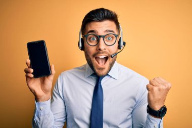 Business operator man with customer service headset from call center showing smartphone screen screaming proud and celebrating victory and success very excited, cheering emotion