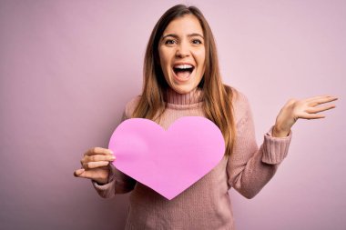 Pembe izole edilmiş arka planda büyük, romantik kağıt kalp şeklinde romantik, sarışın bir kadın. Mutlu ve heyecanlı, kazanan yüz ifadesi zaferi büyük bir gülümsemeyle kutluyor ve ellerini kaldırıyor.
