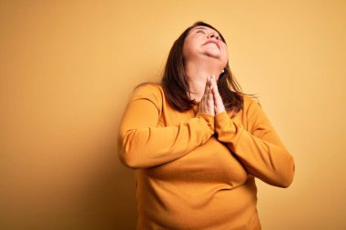 Güzel esmer ve cüsseli bir kadın izole edilmiş sarı arka plan üzerine gündelik süveter giyip elleri bağlı yalvarıyor ve dua ediyor. Yüzünde umut ifadesi var. Çok duygusal ve endişeli. Yalvarıyorum..