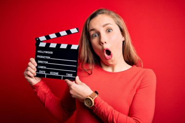 Genç, güzel, sarışın, alkış tutan kadın, kırmızı arka plan üzerinde el çırpma tahtası tutuyor. Şaşırmış bir yüzle korkmuş, korku ifadesinden korkmuş ve heyecanlı.
