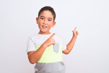 İzole edilmiş beyaz arka plan üzerinde dikilen, gülümseyen ve kameraya bakan, iki eli ve parmağı yandan gösteren güzel çocuk tişörtü giyiyor..