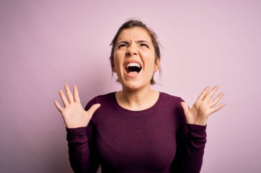 Güzel genç bir kadın pembe izole edilmiş arka planda sıradan bir çörek saç stili takıyor çılgın ve sinirli bir şekilde bağırıyor ve agresif bir ifade ve kollarla bağırıyor. Hayal kırıklığı konsepti.