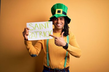Young african american man wearing green hat holding banner celebrating saint patricks day very happy pointing with hand and finger