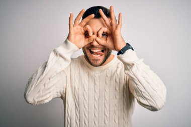 Young handsome man wearing casual sweater standing over isolated white background doing ok gesture like binoculars sticking tongue out, eyes looking through fingers. Crazy expression.
