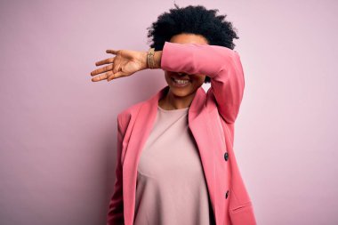 Young beautiful African American afro businesswoman with curly hair wearing pink jacket covering eyes with arm smiling cheerful and funny. Blind concept.