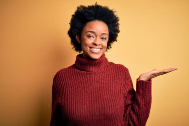 Young beautiful African American afro woman with curly hair wearing casual turtleneck sweater smiling cheerful presenting and pointing with palm of hand looking at the camera.