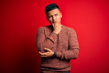 Young handsome latin man wearing casual sweater standing over red background with hand on chin thinking about question, pensive expression. Smiling with thoughtful face. Doubt concept.