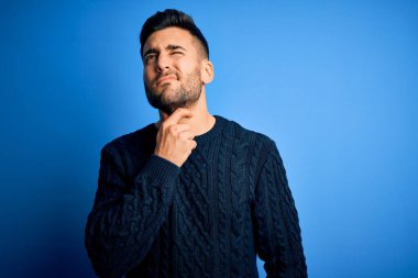 Young handsome man wearing casual sweater standing over isolated blue background Touching painful neck, sore throat for flu, clod and infection