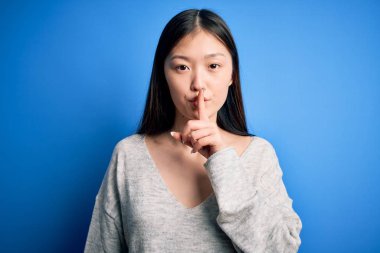 Young beautiful asian woman wearing casual sweater standing over blue isolated background asking to be quiet with finger on lips. Silence and secret concept.