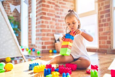 Genç, güzel sarışın kız anaokulunda oyuncaklarla oyun oynuyor, gülümsüyor ve evde yapı taşlarıyla oynuyor.