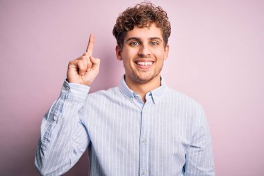 Sarışın, kıvırcık saçlı, beyaz arka planda çizgili gömlek giyen, kendine güvenen ve mutlu bir şekilde gülümseyen genç, yakışıklı bir adam..