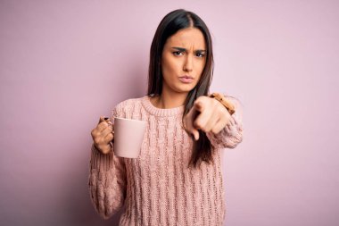 Genç, güzel esmer kadın, izole edilmiş pembe arka planda kahve içerek kamerayı işaret ediyor ve sen, el işareti, ön taraftan pozitif ve kendinden emin bir jest.