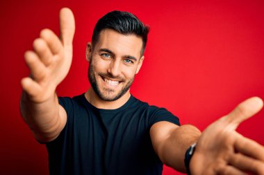Young handsome man wearing casual black t-shirt standing over isolated red background looking at the camera smiling with open arms for hug. Cheerful expression embracing happiness.
