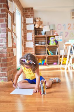Gözlüklü ve tek boynuzlu at diademi takmış güzel bir çocuk. Anaokulunda kağıt ve kalem kullanarak yerde oturuyor.