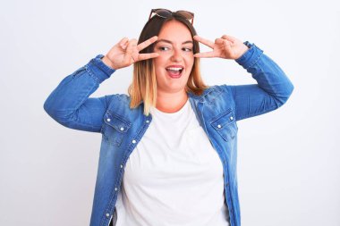 Güzel bir kadın kot gömlek giyiyor, izole edilmiş beyaz arka planda duruyor barış sembolü yapıyor, parmakları yüzünde, neşeyle gülümsüyor zaferi gösteriyor.