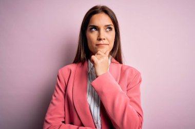 İzole edilmiş pembe arka planda duran ceketli genç ve güzel bir esmer kadın. Elleri çenesinde soru sorarken, düşünceli bir ifade takınıyor. Düşünceli bir yüzle gülümsüyor. Şüphe kavramı.