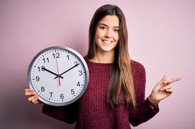 Young beautiful girl doing countdown holding big clock over isolated pink background very happy pointing with hand and finger to the side