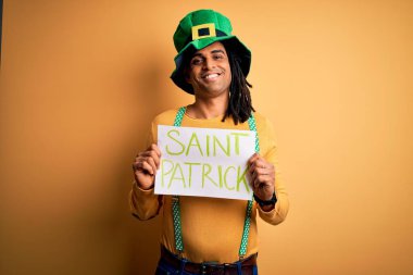 Young african american man wearing green hat holding banner celebrating saint patricks day with a happy face standing and smiling with a confident smile showing teeth