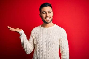 Young handsome man wearing casual white sweater standing over isolated red background smiling cheerful presenting and pointing with palm of hand looking at the camera.