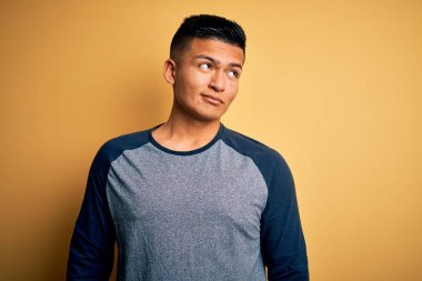 Genç, yakışıklı, günlük tişört giyen, sarı arka planda duran, gülümseyen ve kafasını çeviren Latin bir adam..