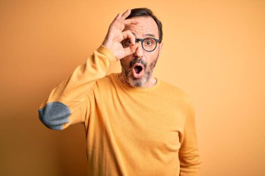Middle age hoary man wearing casual sweater and glasses over isolated yellow background doing ok gesture shocked with surprised face, eye looking through fingers. Unbelieving expression.