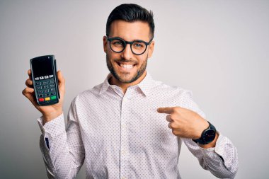 Young handsome man doing payment using dataphone over isolated white background with surprise face pointing finger to himself