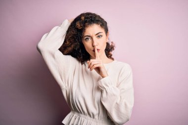Kıvırcık saçlı ve piercingli güzel bir kadın pembe arka planda sıradan bir süveter giyiyor ve parmakları dudaklarında sessiz olmak istiyor. Sessizlik ve gizli konsept.